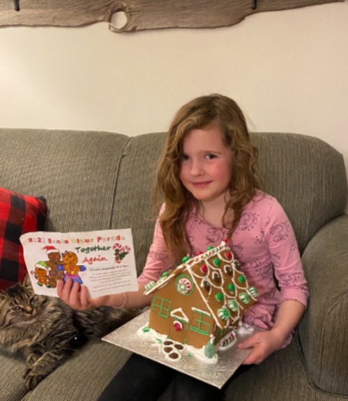 Girl sits on beige couch with long haired cat. Girl holding the her winning entry to the Santa Claus Parade colouring draw and the gingerbread house that she won in the draw. 